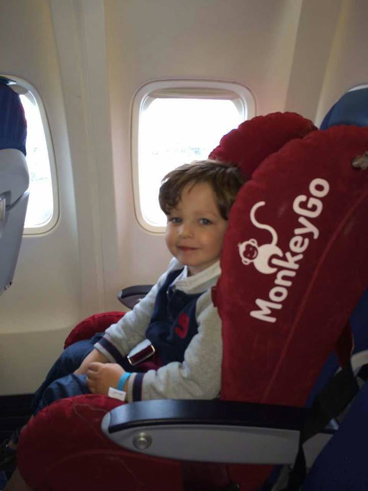 Kind zit zelfstandig in een MonkeyGo™ kinderstoel in een vliegtuig, lacht naar de camera, stevig vastgezet met vliegtuiggordel.